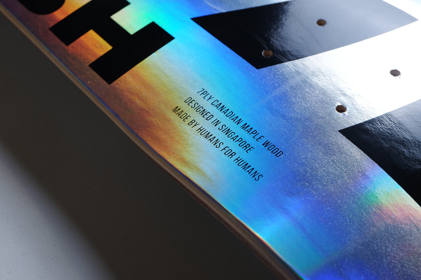 Close-up of a glossy skateboard deck showing part of a black graphic and the underside with a vibrant iridescent rainbow finish and printed text.