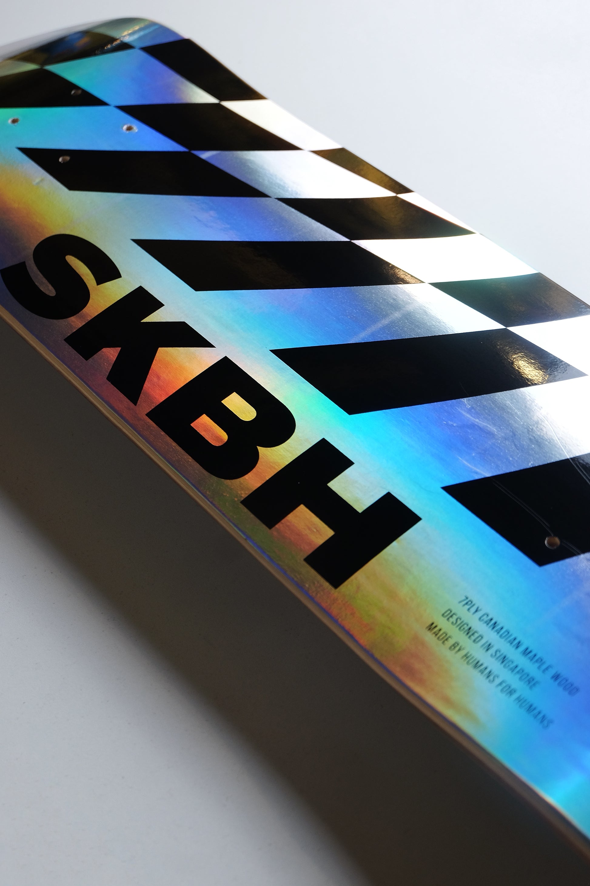 A close-up of a glossy rectangular product with a black-and-white checkered clapperboard design and large black 'SKBH' lettering, showing an iridescent holographic finish on a light gray background.